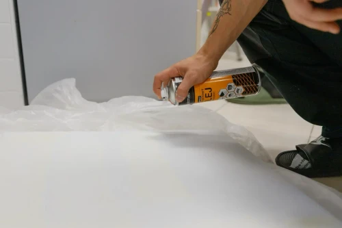 Instalator nanosi klej w sprayu na krawędź białej chmury akustycznej na stanowisku roboczym.